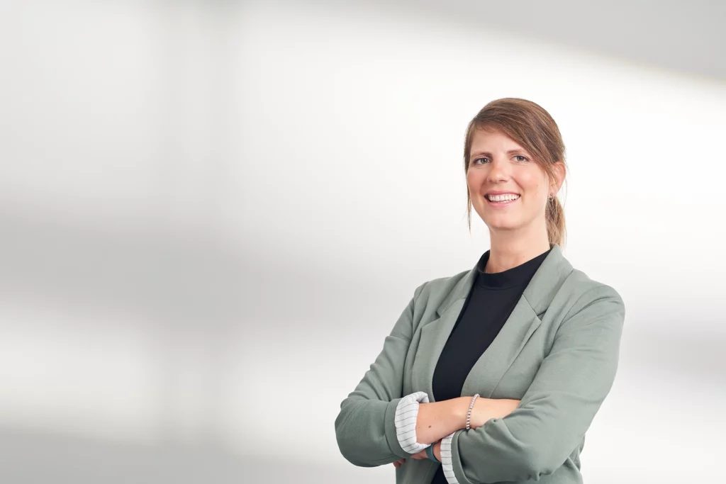 Portrait von Sina Gruhn. Sie steht lächelnd mit verschränkten Armen vor einem hellen, neutralen Hintergrund.