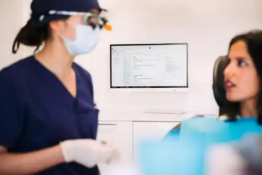 Zu sehen ist eine Szene beim Zahnarzt. Im Fokus ist ein Bildschirm mit Patientendaten. Vorne sieht man unscharf eine Ärztin mit Mundschutz, Handschuhe und Brille auf. Rechts im Bild sitzt eine Patientin die interessiert zuhört.