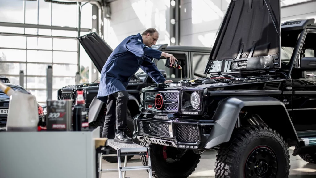 Ein Automechaniker der Öl in den Motor des Tankes eines Brabus gießt. Dabei steht er auf einer kleinen Trittleiter. Im Hintergrund sind noch weitere Autos zu sehen