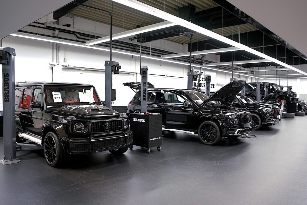 Mehrere Autos der Marke Brabus stehen in einer Werkstatt nebeneinander aufgereiht. Manche dieser Autos haben eine geöffnete Motorhaube. Alle Autos sind schwarz. Es sind verschiedene Automodelle zu sehen